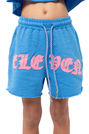 Short Julie Bleu en molleton porté par un enfant, inscription Eleven gothique rose, cordon bicolore et bas effiloché