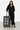 Valise Fugers Gris Foncé M portée par une femme en ensemble noir, patchs colorés et roulettes orange
