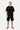 Cardigan Joly Noir à motifs brodés porté par un mannequin homme blond avec short noir assorti, baskets roses, vue en pied