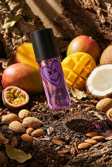 Brume parfumée Glow Pop violette posée sur de la terre entourée de mangues, noix de coco, fruits de la passion et amandes