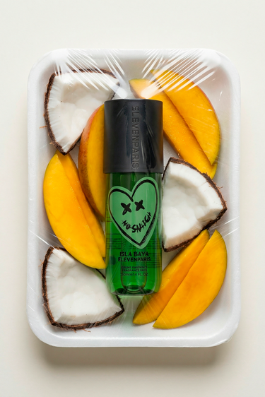 Parfum Isla Baya Eleven Paris, flacon vert au cœur souriant posé sur des morceaux de mangue et de noix de coco
