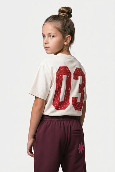 T-shirt Tiami Écru porté par une enfant, de dos, grand numéro 03 rouge, avec pantalon bordeaux